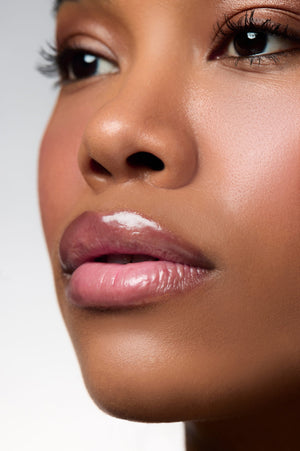 Close-up of a woman's face showing glossy pink lips with a white cosmetic product applied on the upper lip, against a light gray background.