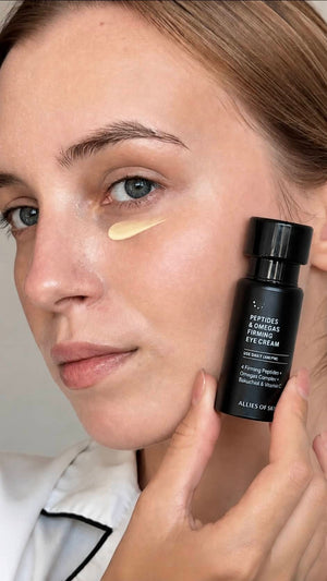 Close-up of a woman applying Peptides & Cinnabar Firming Eye Cream from Allies of Skin, holding the black bottle next to her eye where a smear of the cream is visible on her cheek.